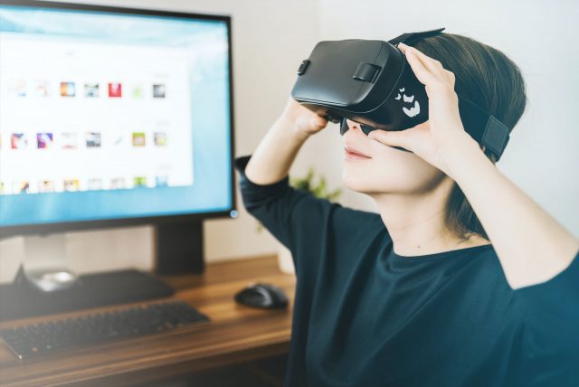 Teaserbild der Computerspielschule. Ein Kind trägt eine VR-Brille mit Logo der Computerspielschule und schaut nach oben. Im Hintergrund befindet sich ein Schreibtisch mit einem Computer.