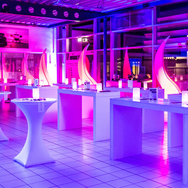 Teaserbild First Contact. Stehtische sind in einem rosa-blau beleuchteten Ausstellungsraum aufgestellt.