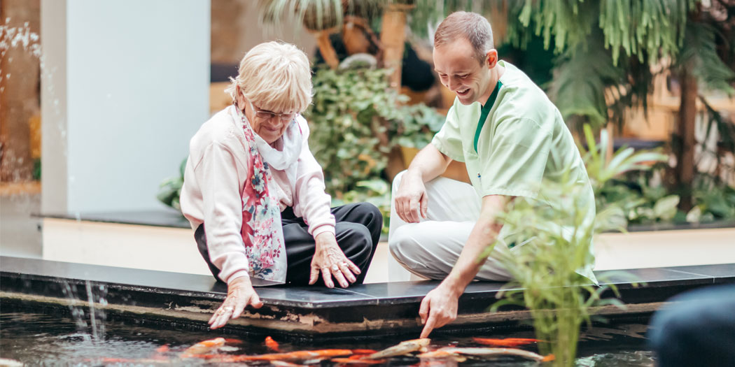 Ein Mitarbeiter der Stiftung Seniorenhilfe Zeitz sitzt mit einer Seniorin an einem Teich mit Fischen.