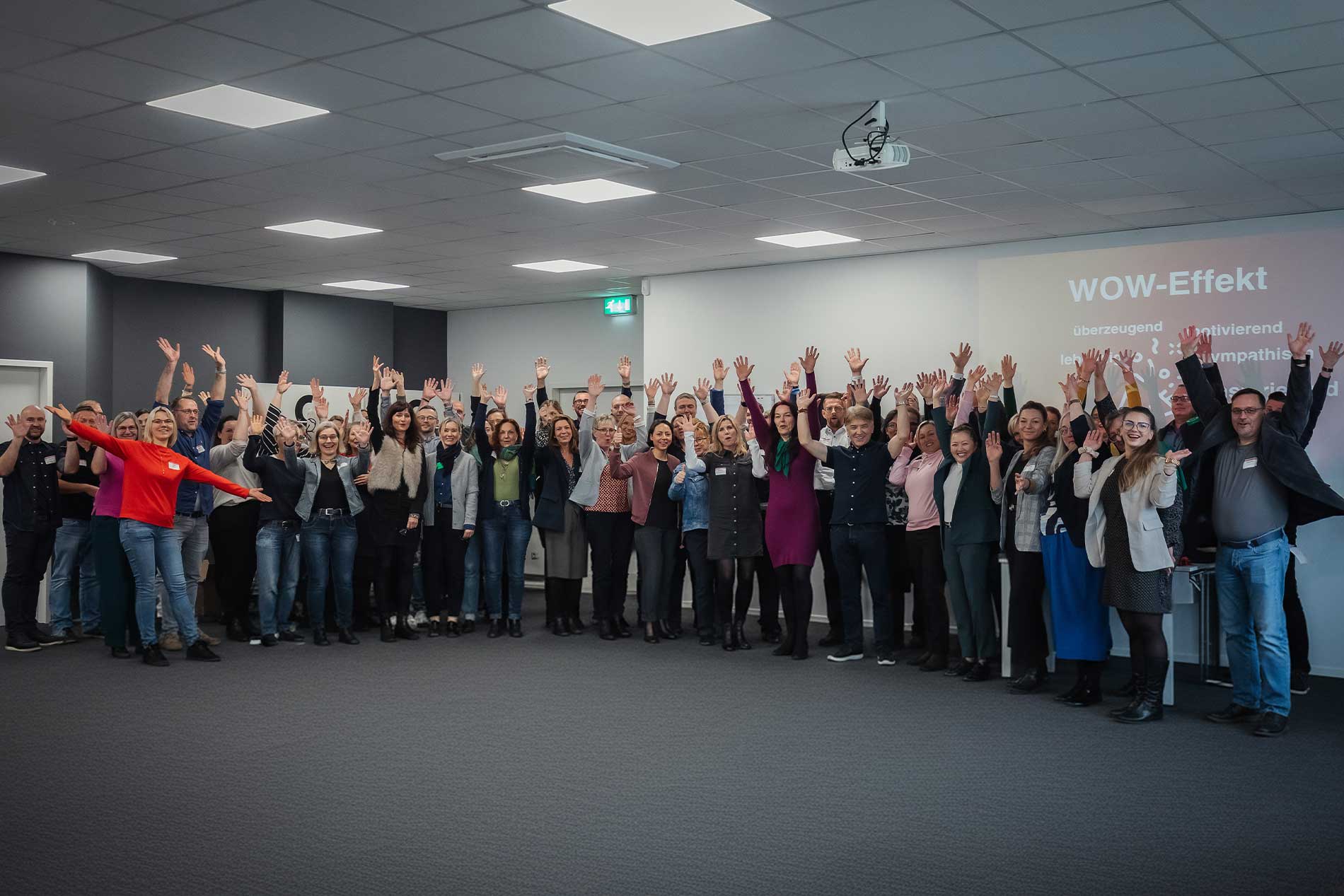 Gruppenfoto eines KI-Workshops mit Phuong Tran: Eine Gruppe von Teilnehmenden steht in einem Seminarraum und hebt lächelnd die Hände. Im Hintergrund ist eine Präsentationsfolie mit dem Begriff „WOW-Effekt“ zu sehen. Das Bild zeigt die positive, aktivierende Atmosphäre eines Workshops zur KI-Strategie und KI-Umsetzung.