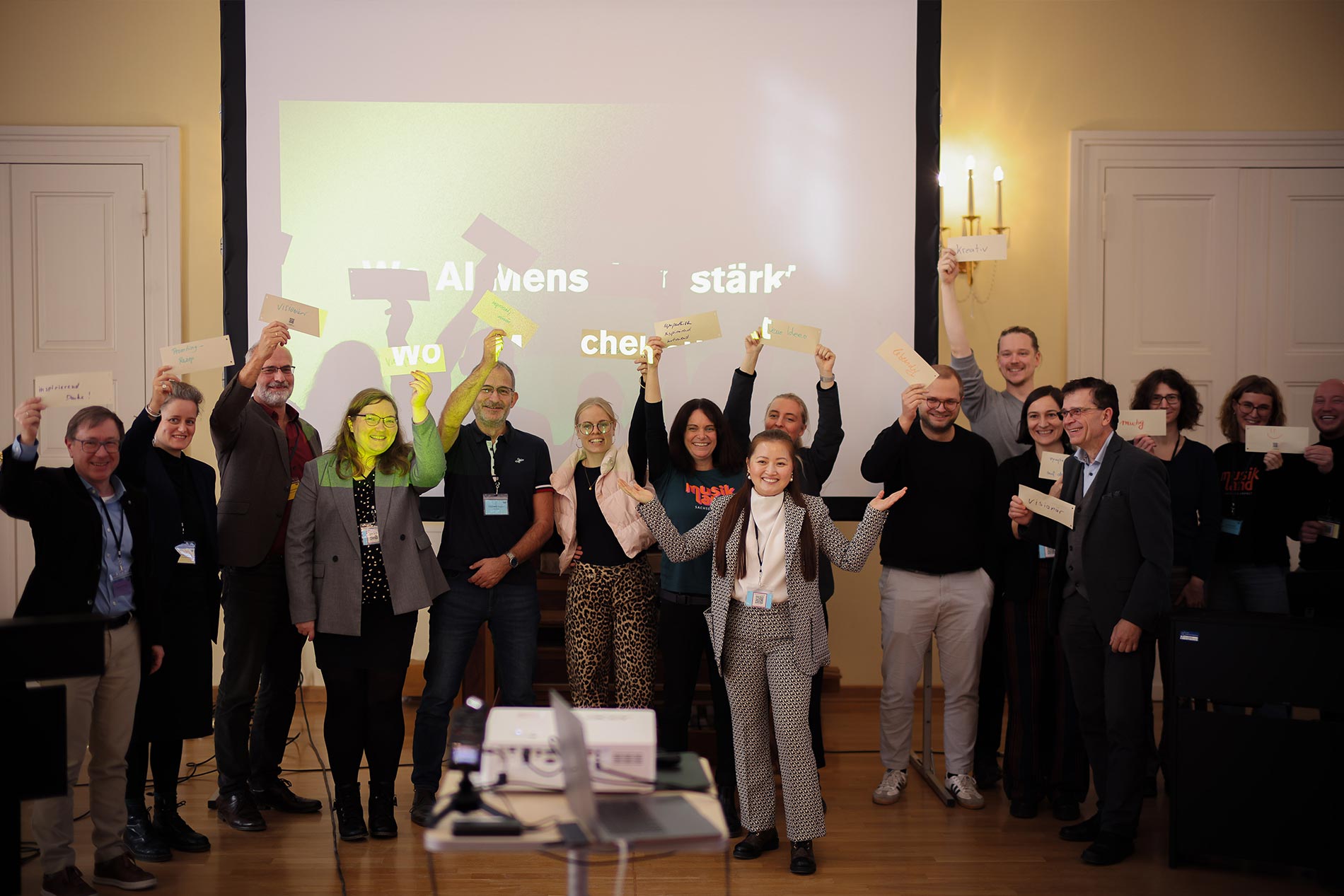 Phuong Tran steht mit einer Workshop-Gruppe in Magdeburg vor einer Präsentationsfläche. Die Teilnehmenden halten beschriftete Karten hoch und lächeln in die Kamera. Das Bild zeigt eine aktive, gemeinschaftliche Workshop-Situation zum Thema Künstliche Intelligenz und kreative Zusammenarbeit.
