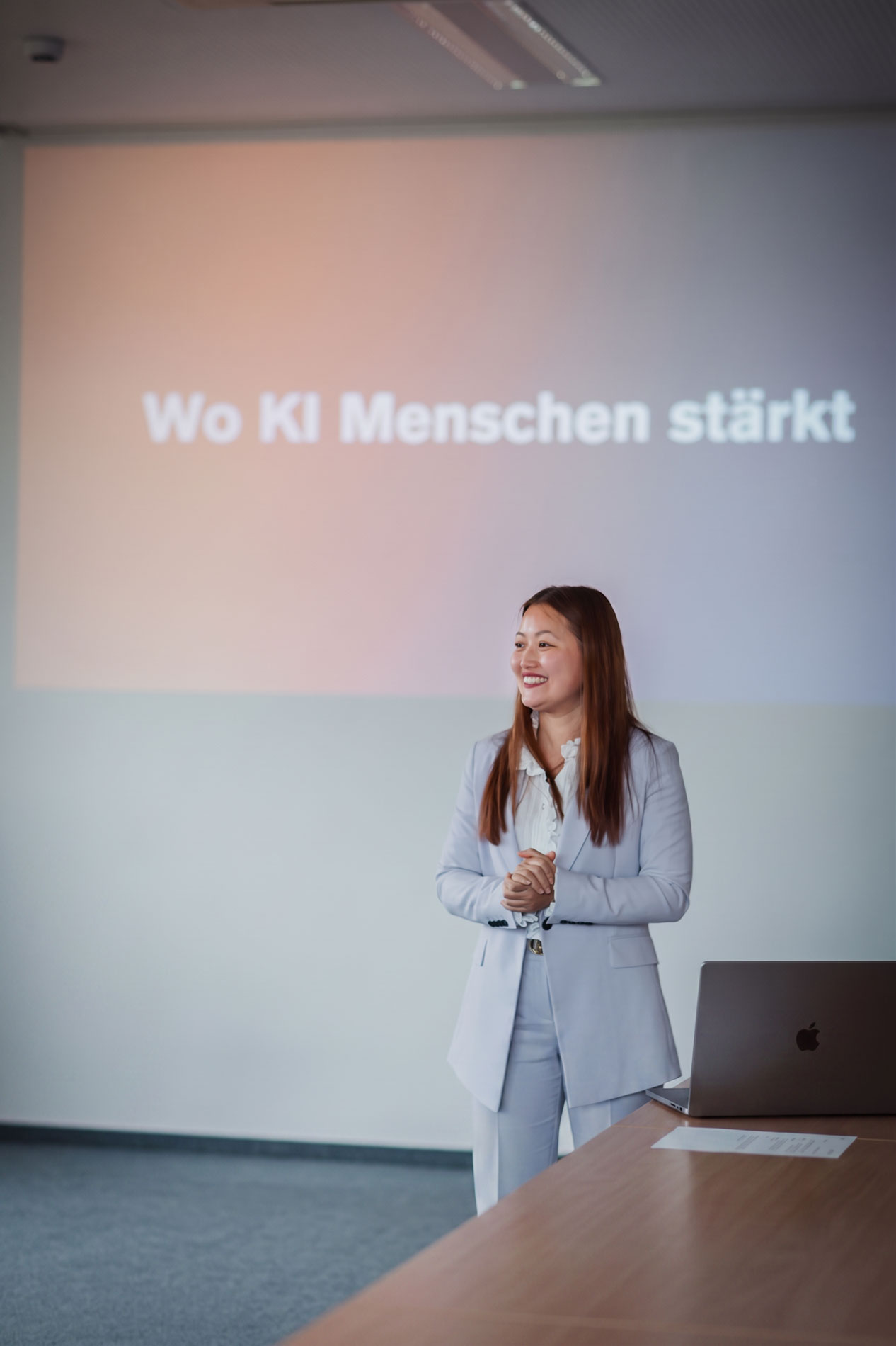 **Alt-Text:** Phuong Tran steht lächelnd in einem Seminarraum vor einer Präsentationswand mit der Aufschrift „Wo KI Menschen stärkt“. Neben ihr befindet sich ein Laptop auf einem Tisch. Das Bild zeigt eine professionelle, positive Vortragssituation zum menschzentrierten Einsatz von Künstlicher Intelligenz.